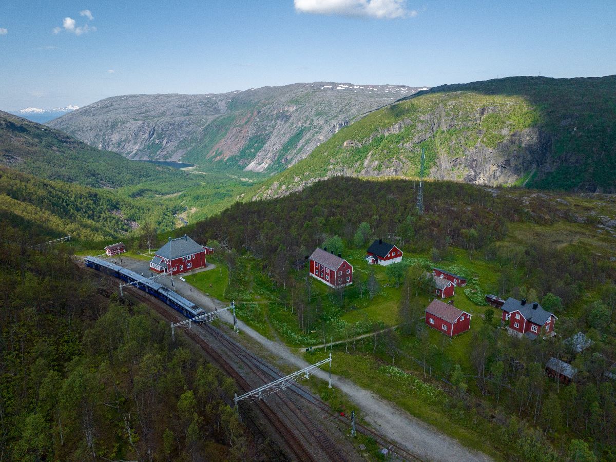 Luftfoto av tog på jernbane ved røde hus i grønn fjelldal, med bratte fjellsider og blå himmel..
