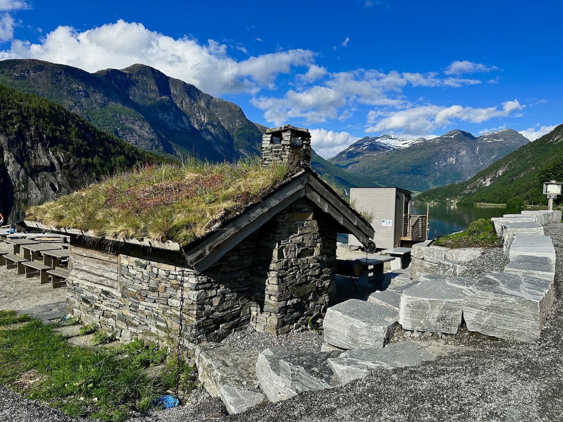 Small stone cabin with a grass roof and chimney beside a fjord, backed by green mountains under blue sky..
