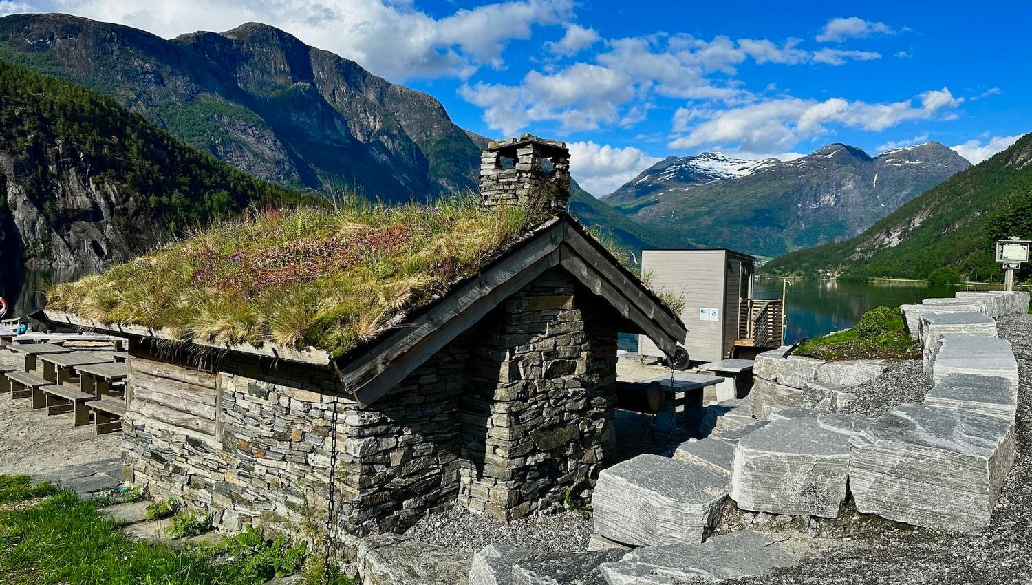 Small stone cabin with a grass roof and chimney beside a fjord, backed by green mountains under blue sky..