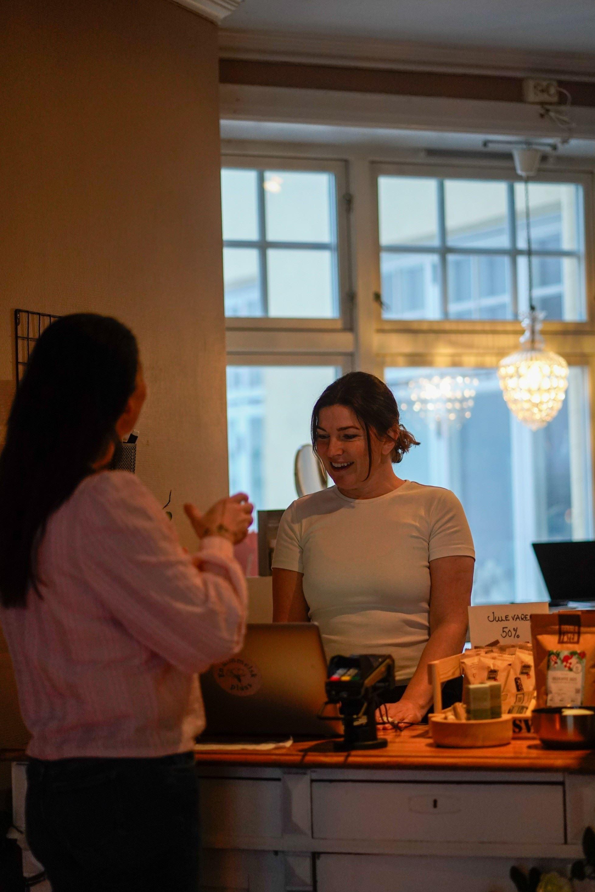 Two women talk at a shop counter with a laptop, card reader, and packaged goods under warm pendant lights..