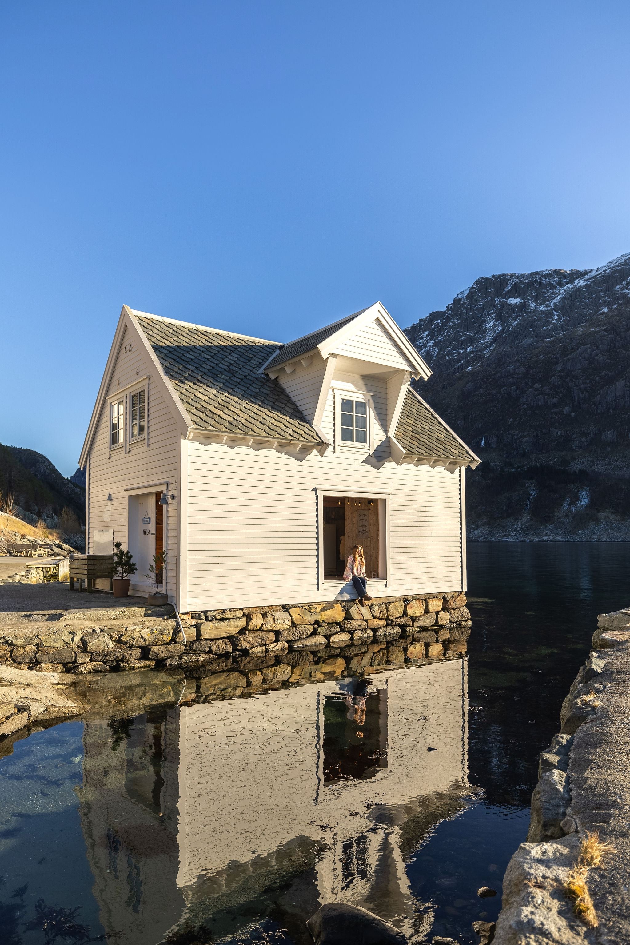 A white wooden boathouse sits on a stone pier beside calm water, reflecting the building and nearby mountains at the Seahouse at Hornelen Panorama in Bremanger...