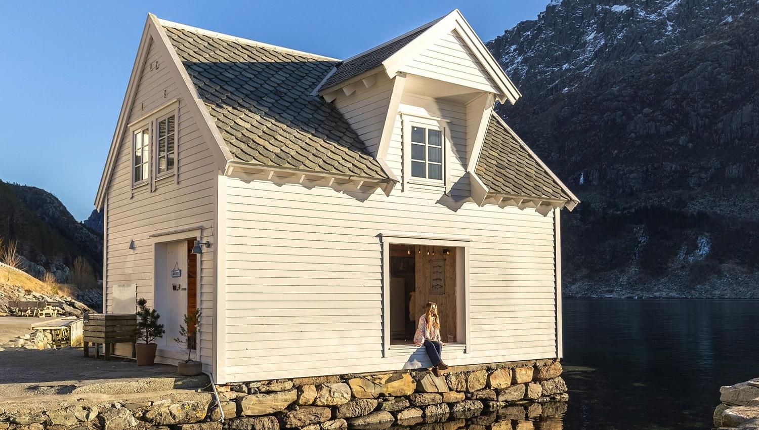 A white wooden boathouse sits on a stone pier beside calm water, reflecting the building and nearby mountains at the Seahouse at Hornelen Panorama in Bremanger...