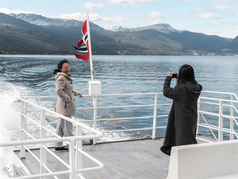 To passasjerar tar selfie på akterdekket under fjordcruise på Hardangerfjorden, snødekte fjell i bakgrunnen.