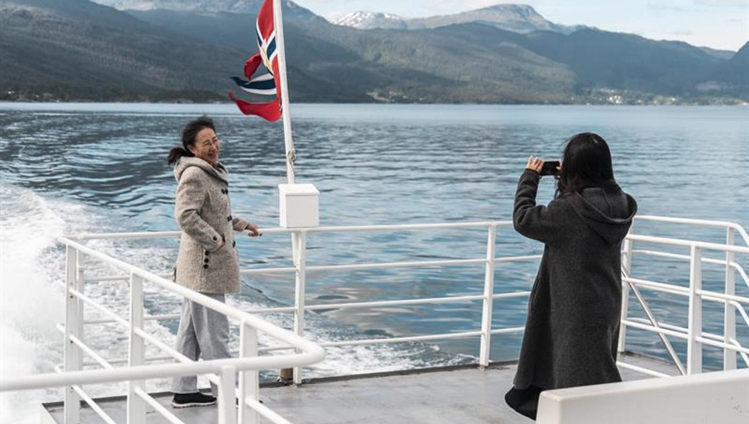 To passasjerar tar selfie på akterdekket under fjordcruise på Hardangerfjorden, snødekte fjell i bakgrunnen.