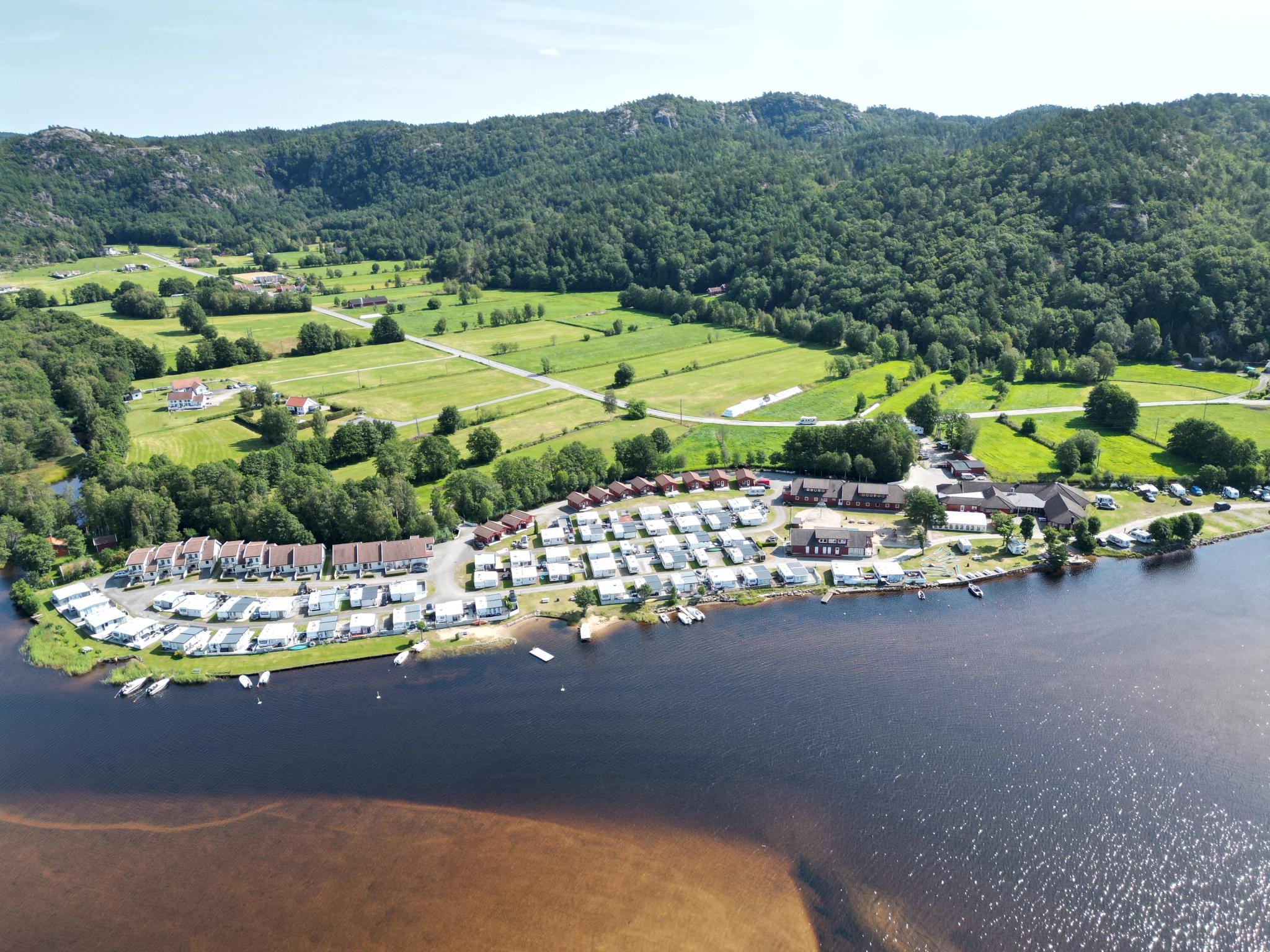 Oversiktsbilde av Solstrand Camping, mange campingvogner og hytter, er langs vannet, bak campingplassen er det store jorder og skog bak det igjen. blå himmel