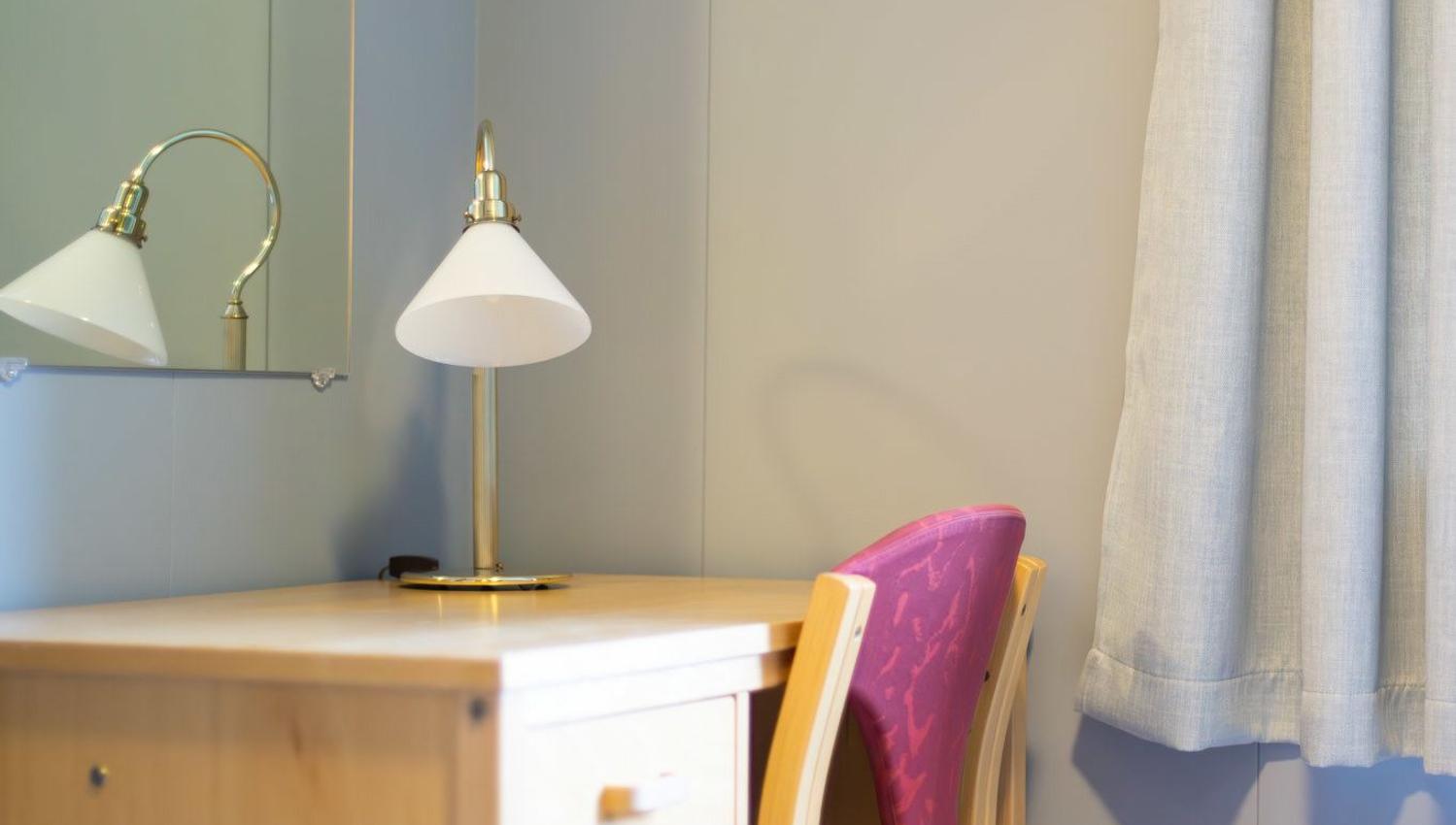 Wooden desk with a brass-and-white lamp, mirror, and a purple chair beside a window with light curtains..