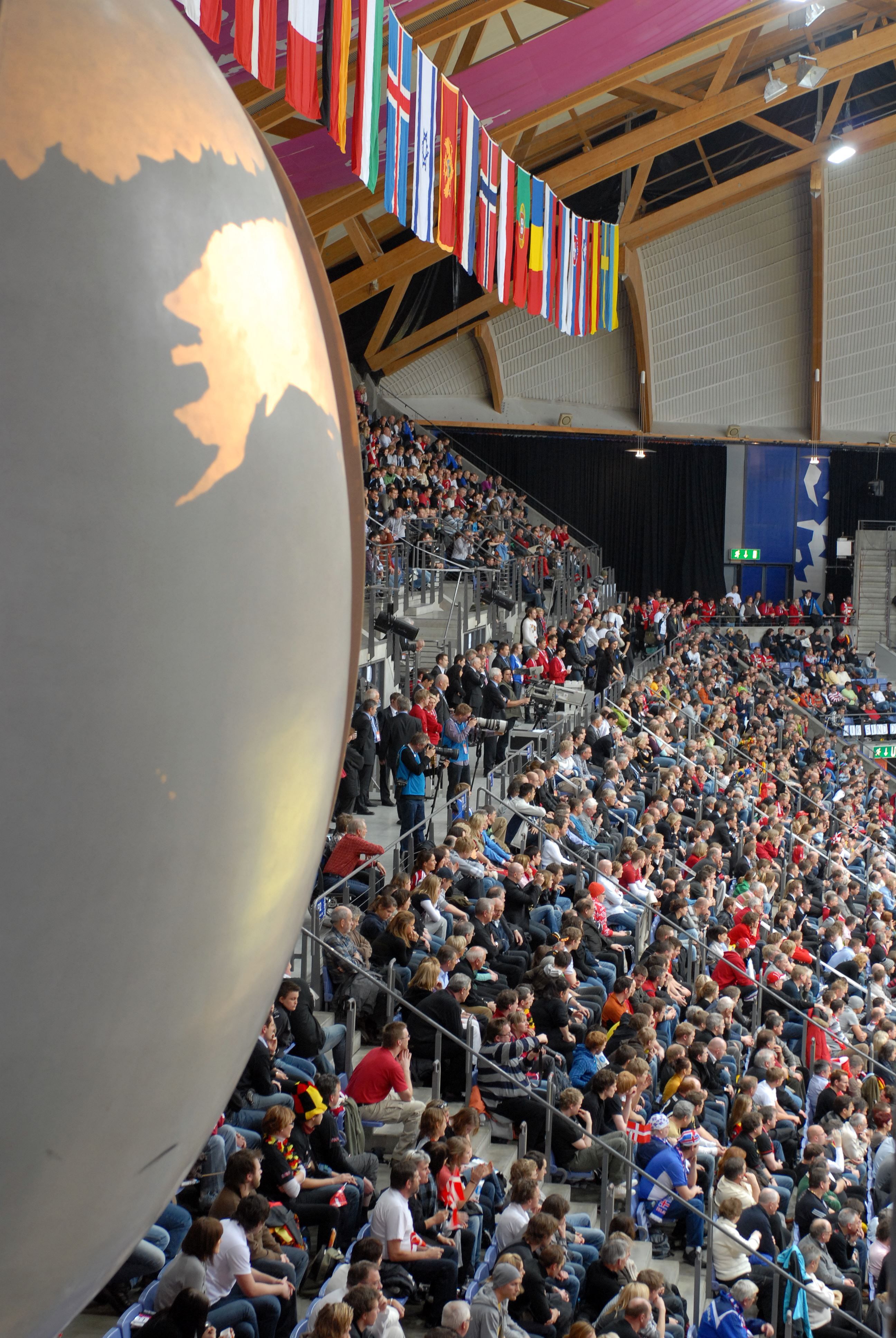 Tettpakket tribune i en innendørsarena med hengende flaggrekke i taket og en stor globus til venstre i bildet..