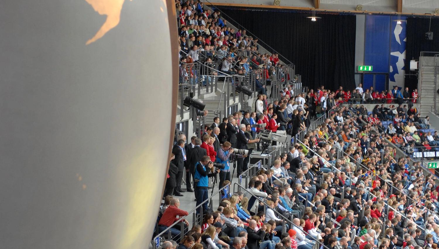 Tettpakket tribune i en innendørsarena med hengende flaggrekke i taket og en stor globus til venstre i bildet..