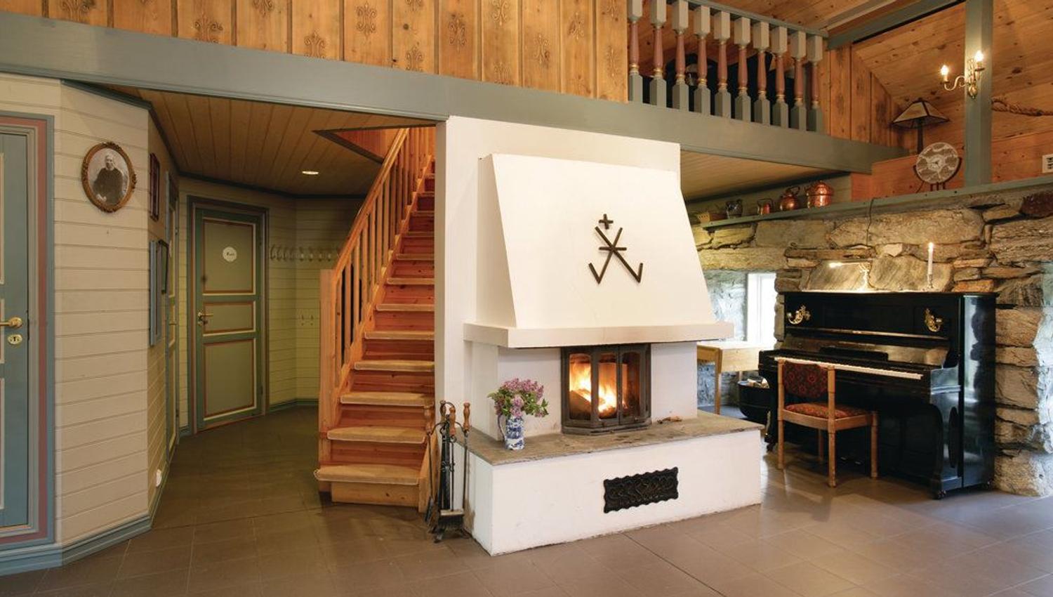 Orheimstunet in Nordfjord - Rustic cabin interior with a central white fireplace, wooden stairs to a loft, stone wall, and a black piano..