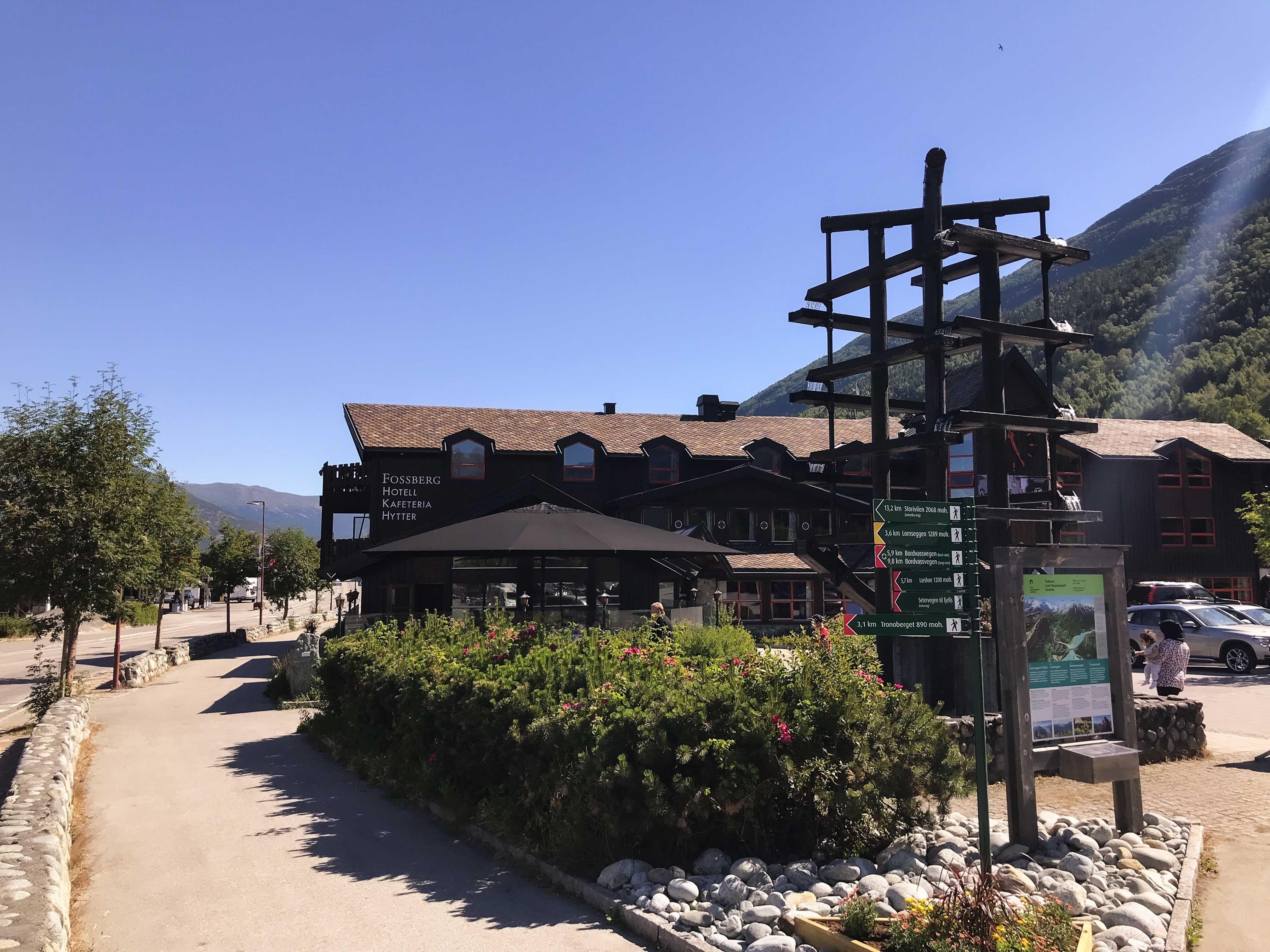 A wooden lodge beside a paved path with shrubs, a tall signpost and map board, and forested mountains under a clear blue sky..