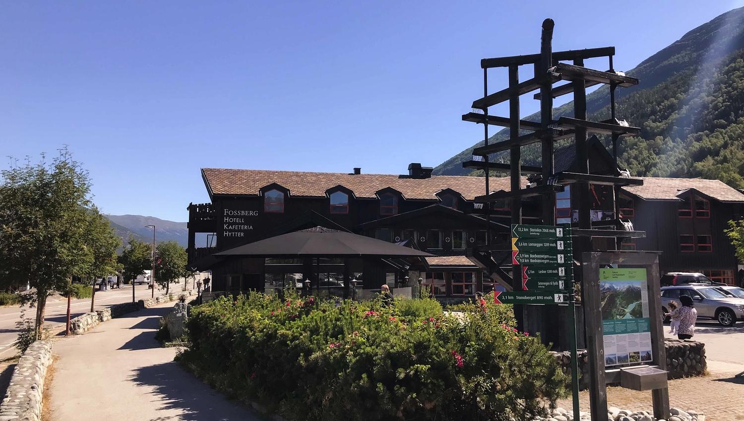 A wooden lodge beside a paved path with shrubs, a tall signpost and map board, and forested mountains under a clear blue sky..
