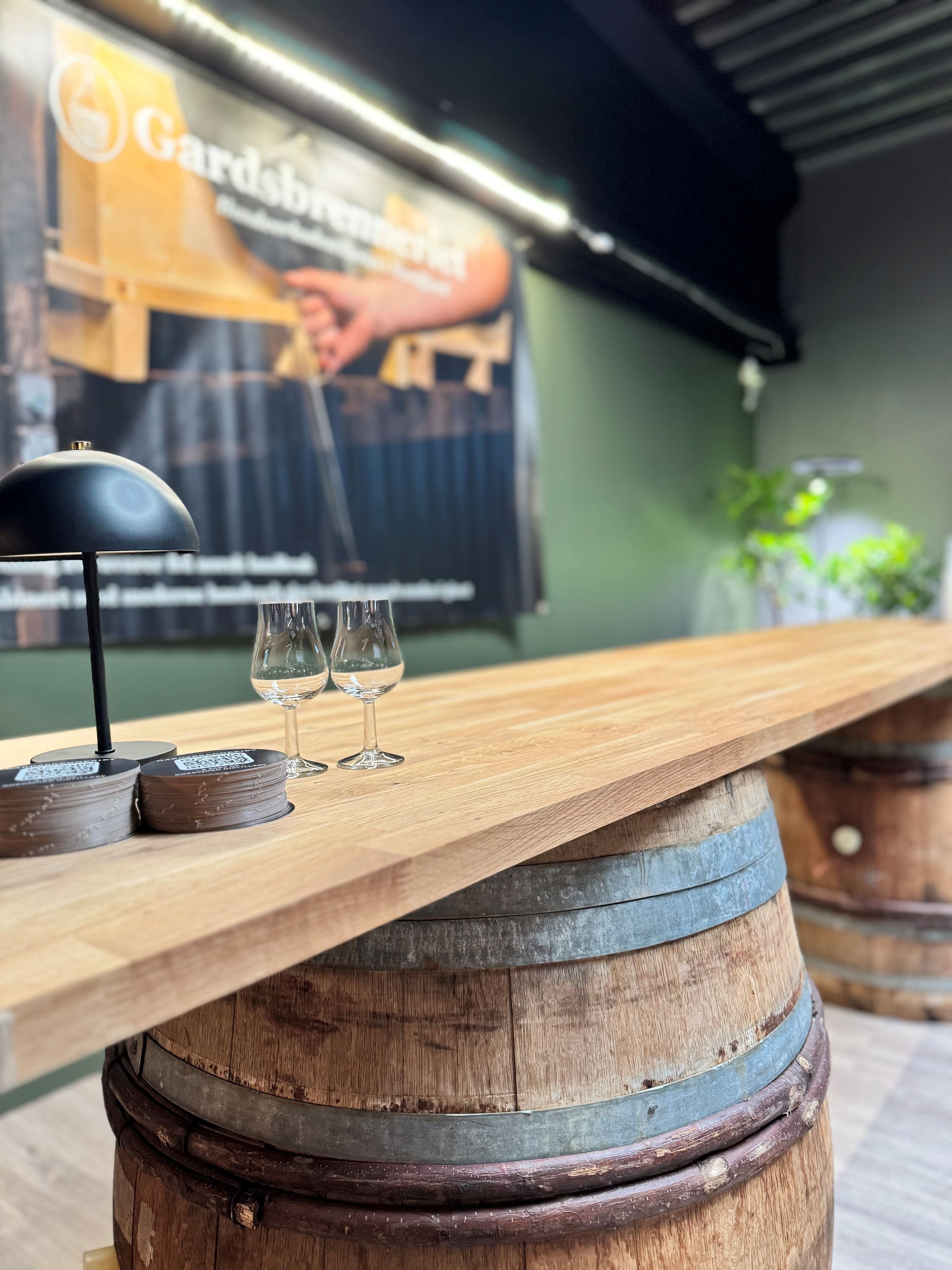 Wooden bar counter on a whiskey barrel base with two tasting glasses and a small lamp in a rustic room.