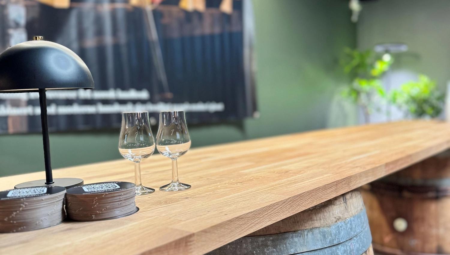 Wooden bar counter on a whiskey barrel base with two tasting glasses and a small lamp in a rustic room.