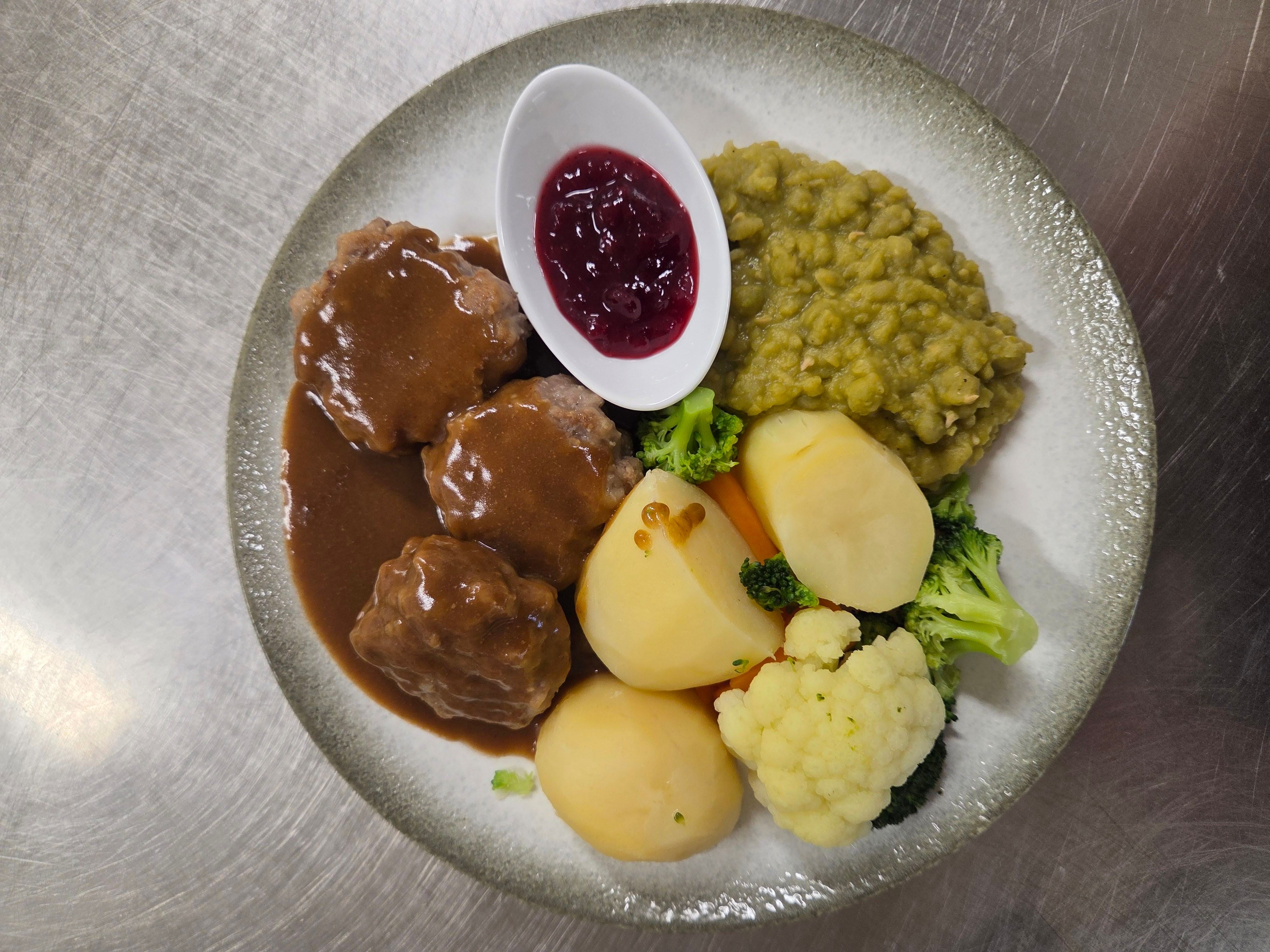 Overhead view of a plate with meatballs in brown gravy, potatoes, mixed vegetables, peas, and lingonberry sauce.