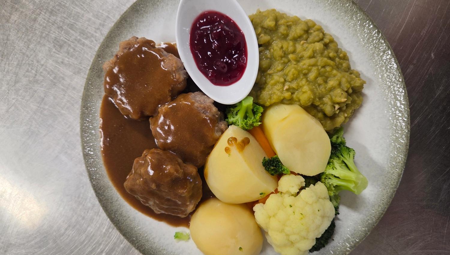 Overhead view of a plate with meatballs in brown gravy, potatoes, mixed vegetables, peas, and lingonberry sauce.