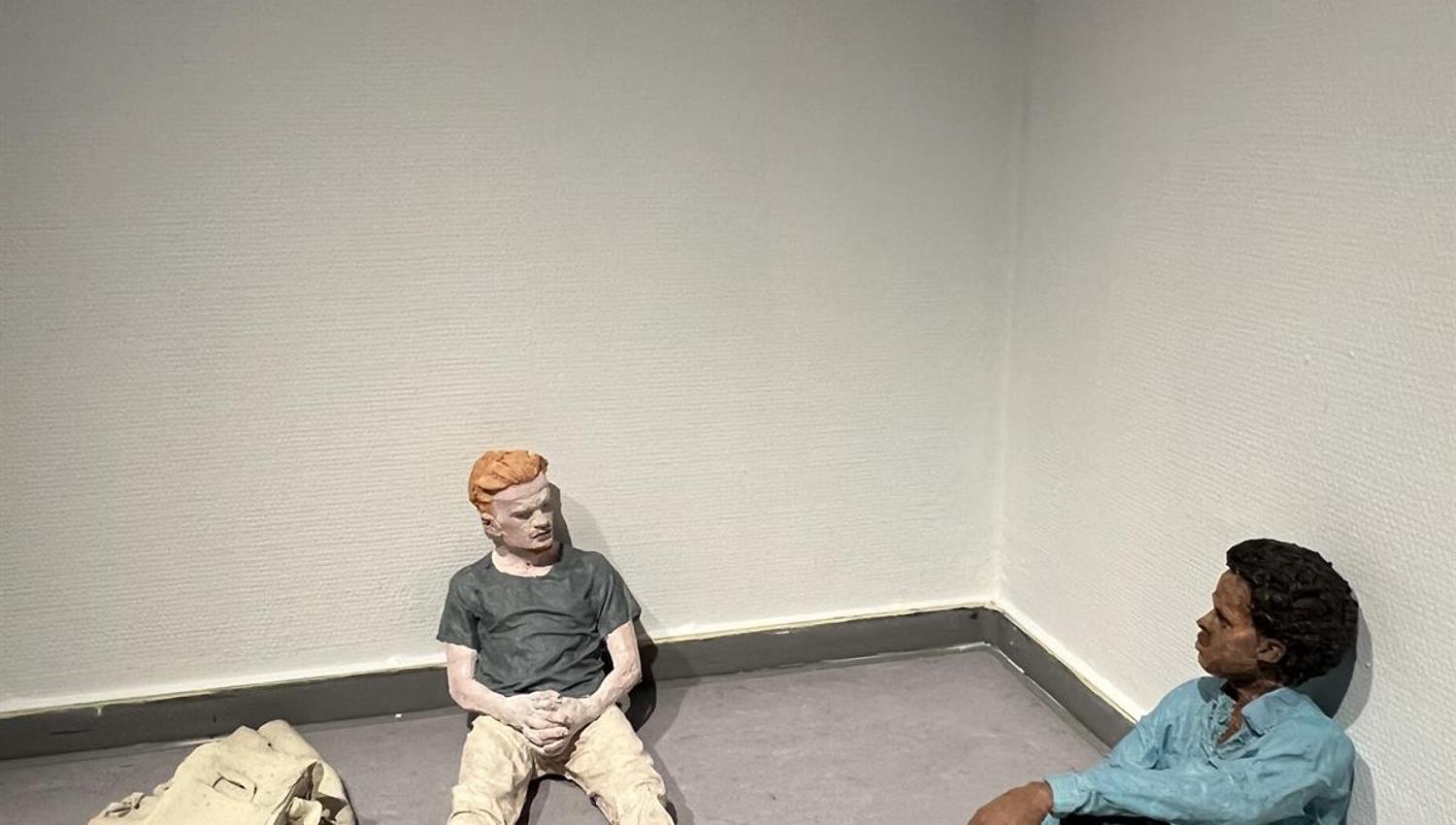 Two sculptural figures sitting on the floor near a wall with objects placed around.