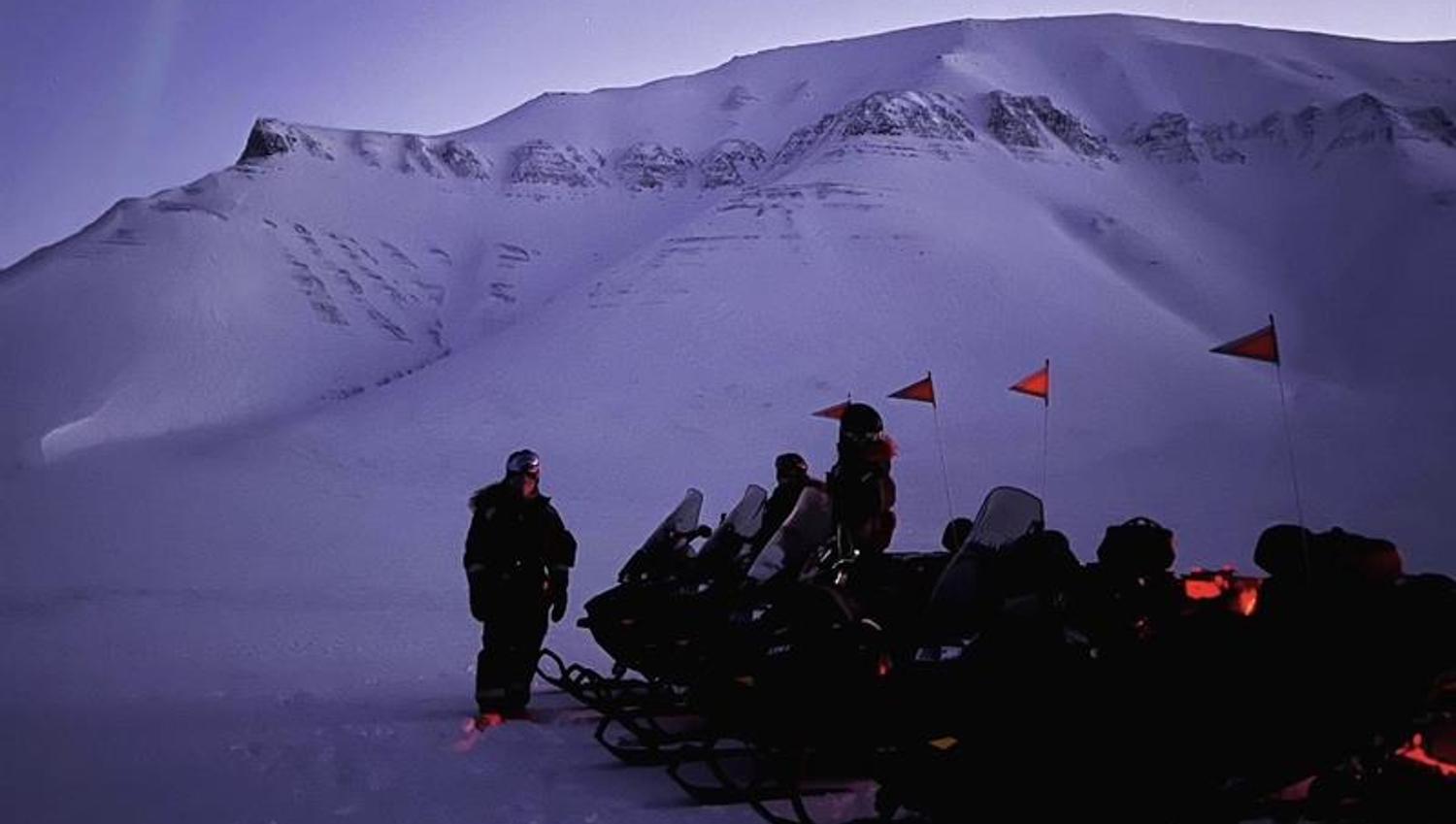 Fire snøscootere står parkert på en slette med et stort fjell i bakgrunnen. Nordlys på himmelen