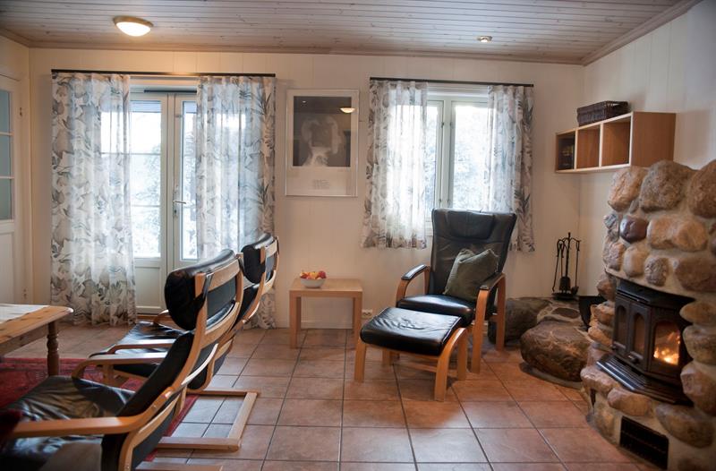 Stue på Hillestad Feriesenter med fyr på peisen og godstol i svart lær, oransje fliser på gulvet, vindu og terrasse dør med vinduer og blomstrete gardiner.
