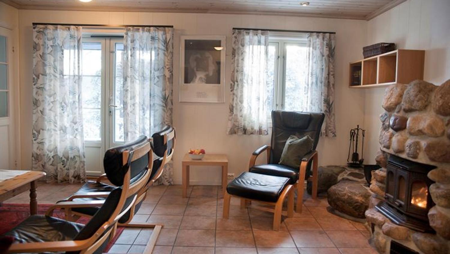 Stue på Hillestad Feriesenter med fyr på peisen og godstol i svart lær, oransje fliser på gulvet, vindu og terrasse dør med vinduer og blomstrete gardiner.