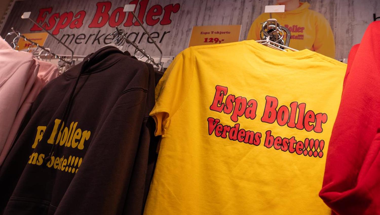 A yellow T-shirt with printed text hanging on a clothing rack in a store, surrounded by other garments and promotional items.