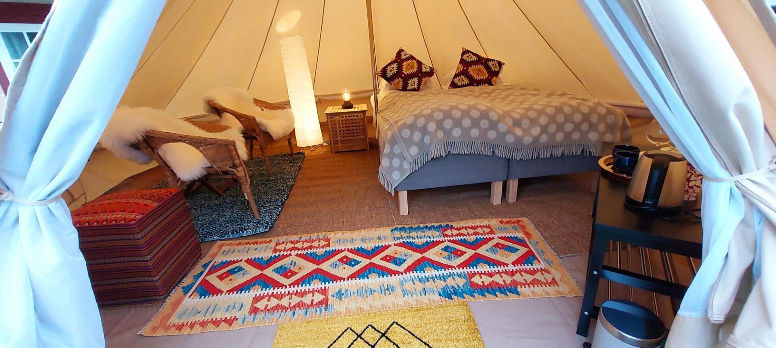 Interior of a glamping tent with a bed, rugs, and a small seating area, decorated with warm colors and textiles.
