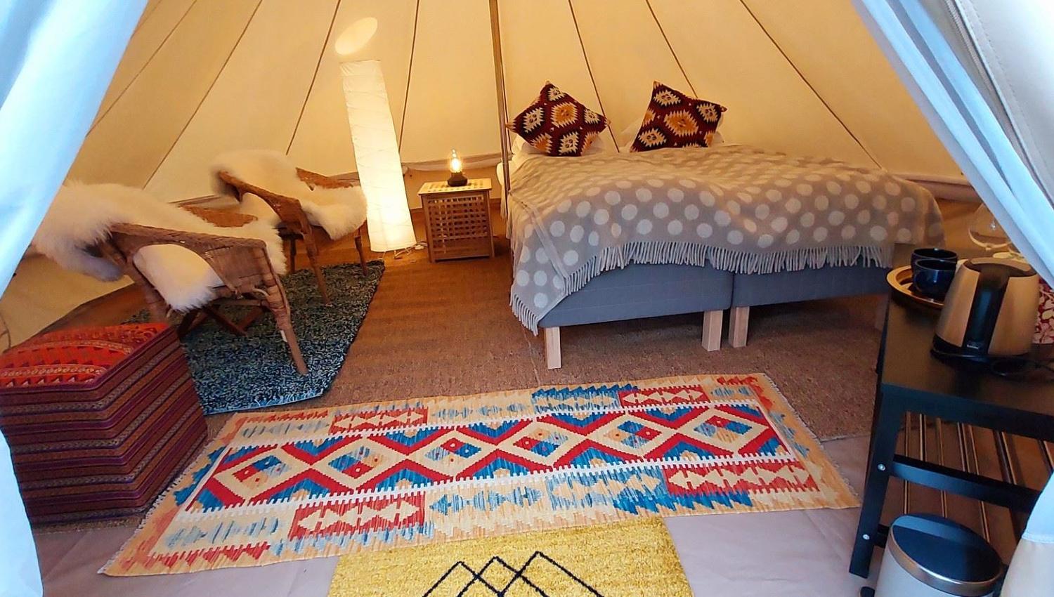 Interior of a glamping tent with a bed, rugs, and a small seating area, decorated with warm colors and textiles.