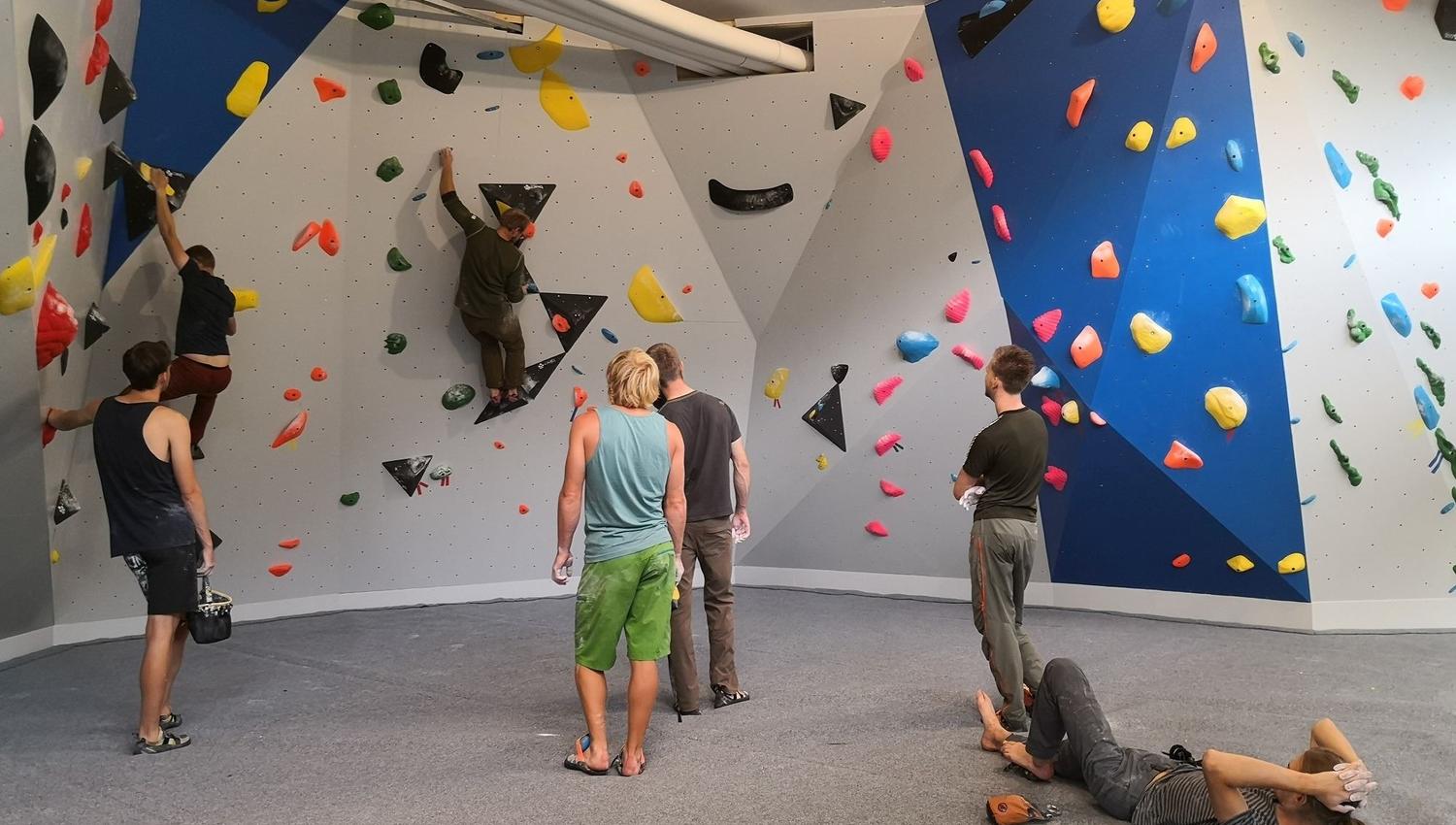 Indoor bouldering wall with colorful holds as several climbers practice, watch, and rest on padded floor..