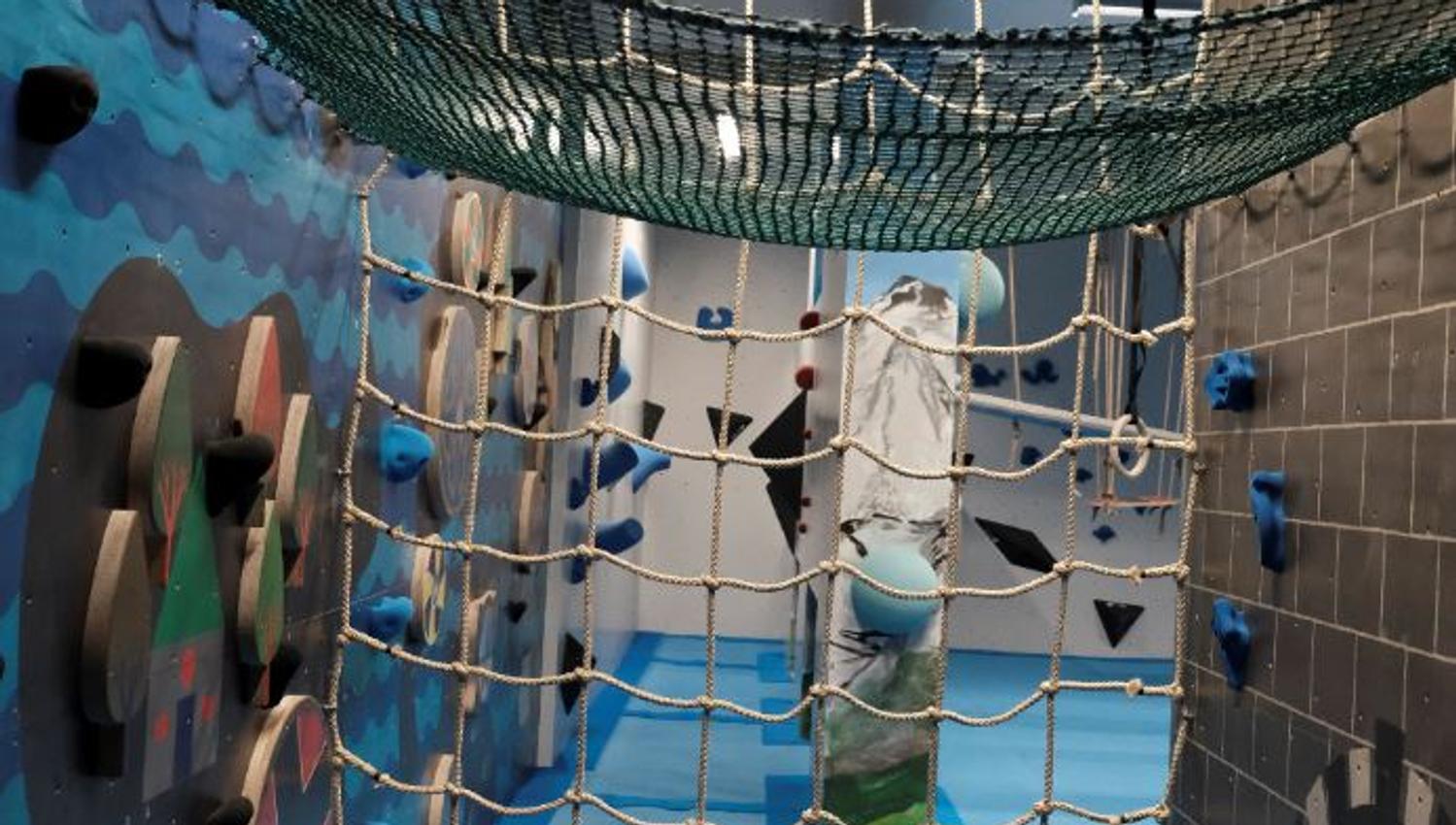 Indoor play area with rope nets, climbing holds on walls, blue padded floor, and a hanging hammock net overhead..