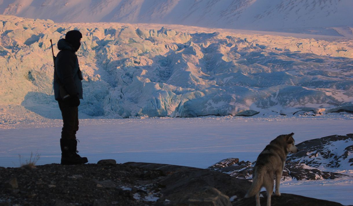 A person with a rifle on their back and a husky in the foreground, with a frozen fjord and a massive glacier in the background