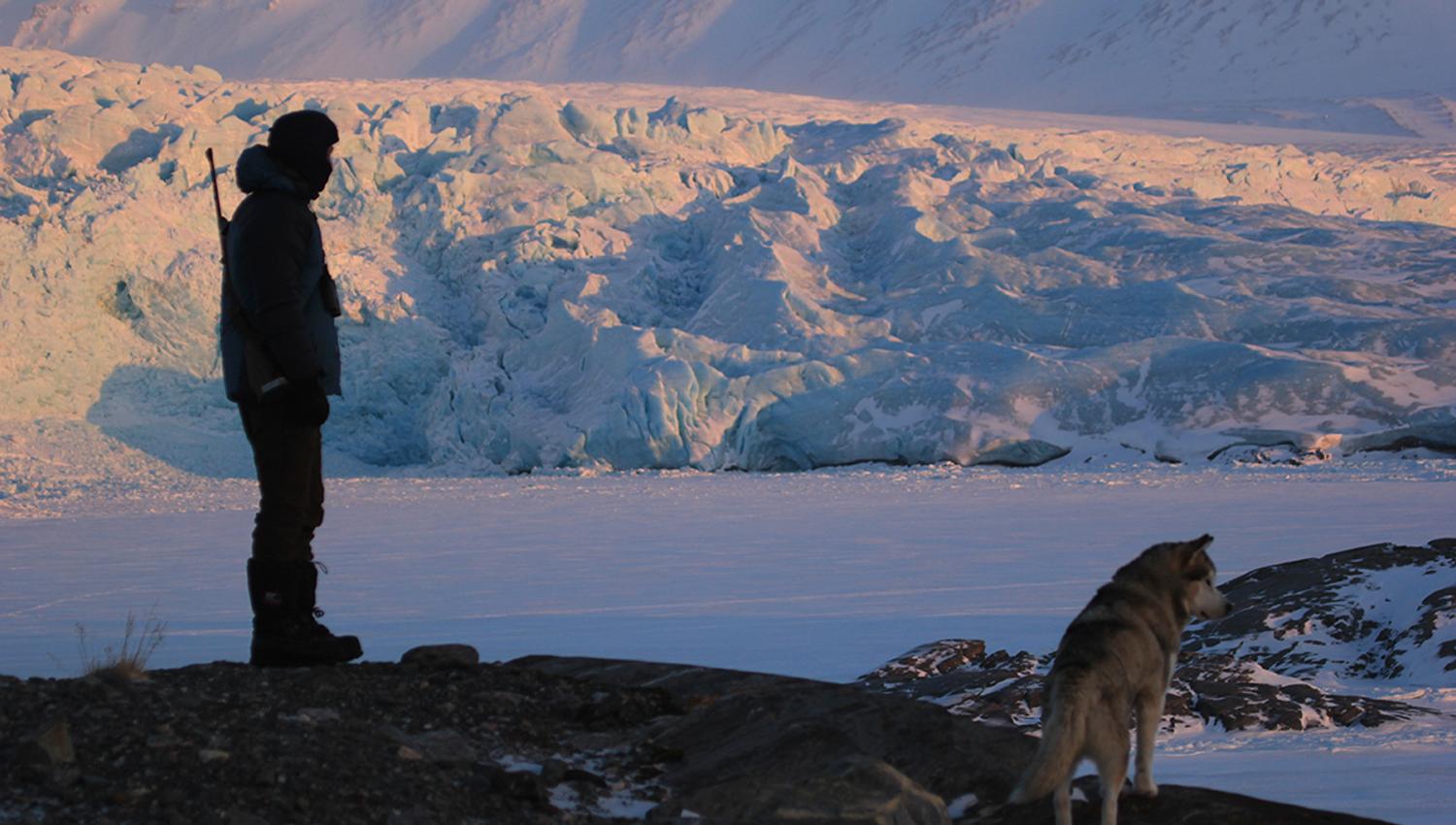 A person with a rifle on their back and a husky in the foreground, with a frozen fjord and a massive glacier in the background