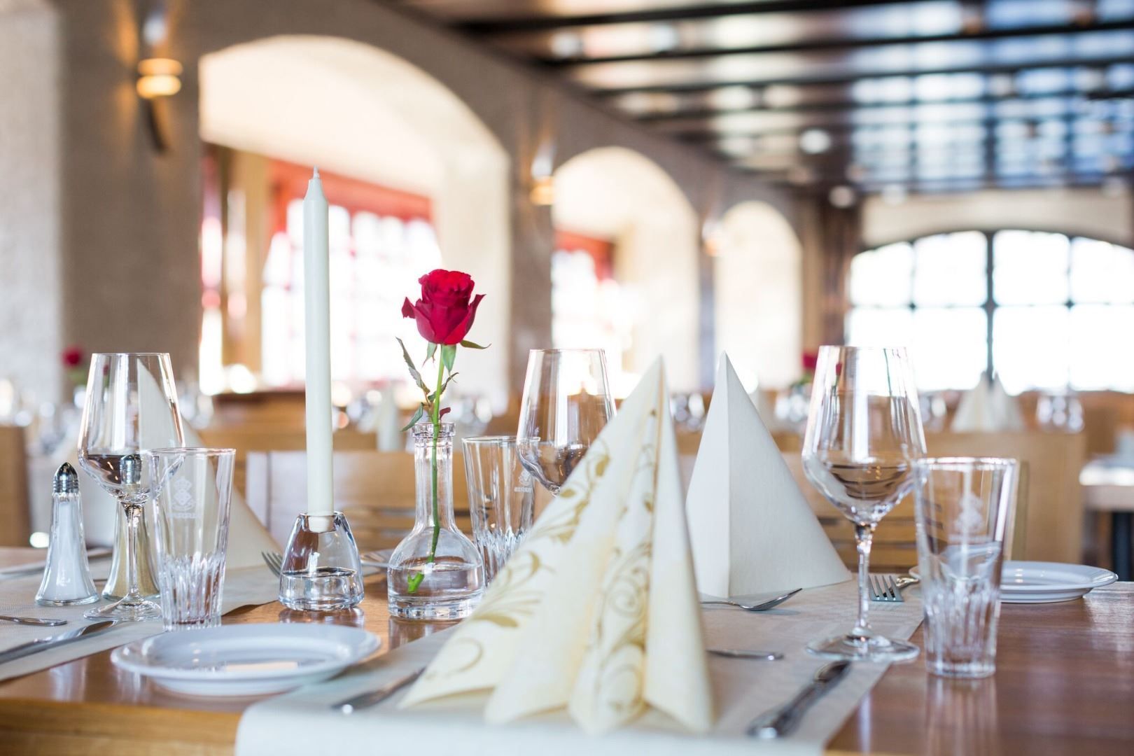 Dekka bord i restaurant med glas, bestikk, serviettar, lysestake og ei raud rose i vase, mjukt dagslys..