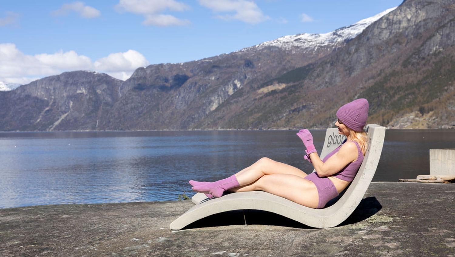 A woman dressed in a purple winter bikini, gloves and a hat, sitting in one of the beton chairs at Kråkeskarvet beach.