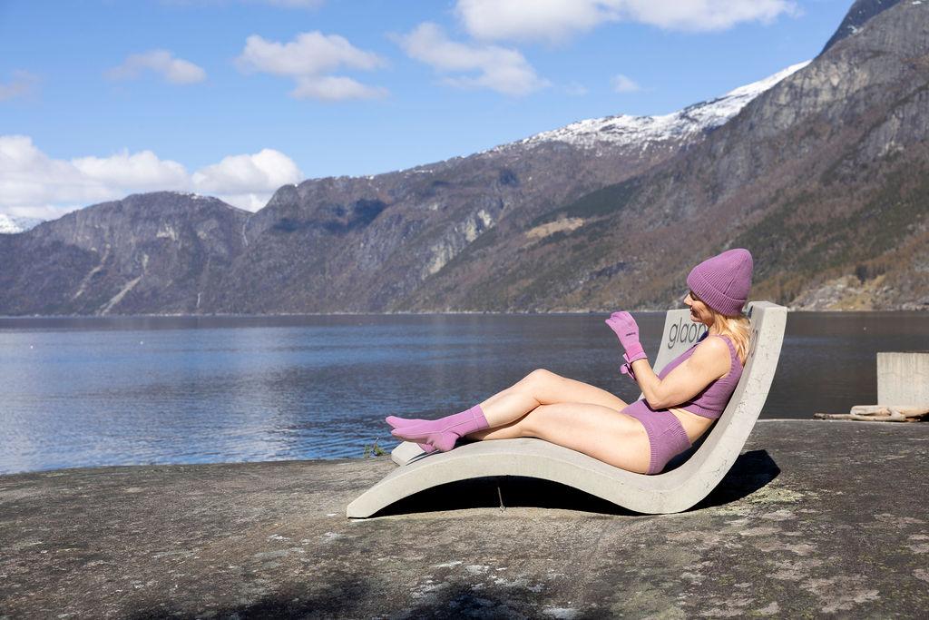 A woman dressed in a purple winter bikini, gloves and a hat, sitting in one of the beton chairs at Kråkeskarvet beach.