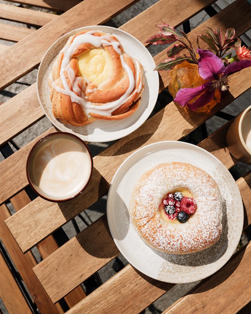 Coffee cups and freshly baked pastries are served on a wooden outdoor table at Iversen Bakery and Gallery, with flowers and sunlight creating a cosy c