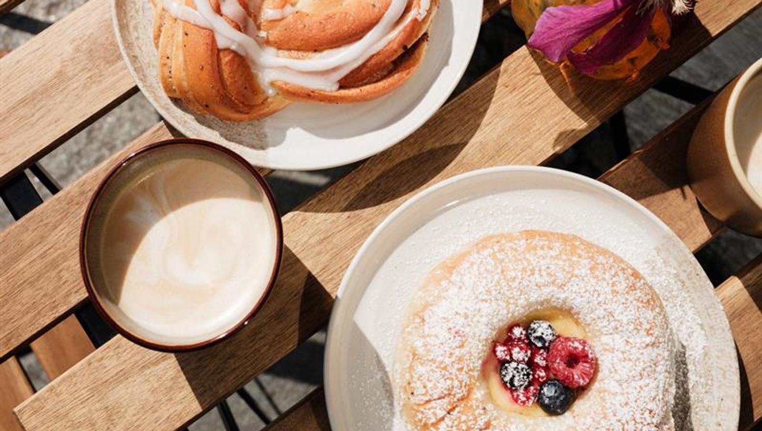 Coffee cups and freshly baked pastries are served on a wooden outdoor table at Iversen Bakery and Gallery, with flowers and sunlight creating a cosy c