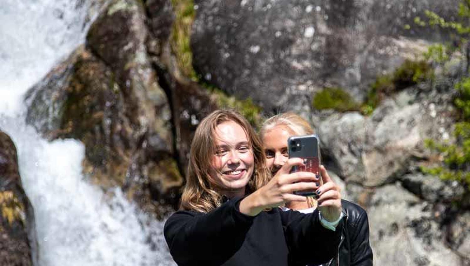 To unge kvinner tar selfie framfor ein foss i Hardanger under eit fjordcruise-stopp.