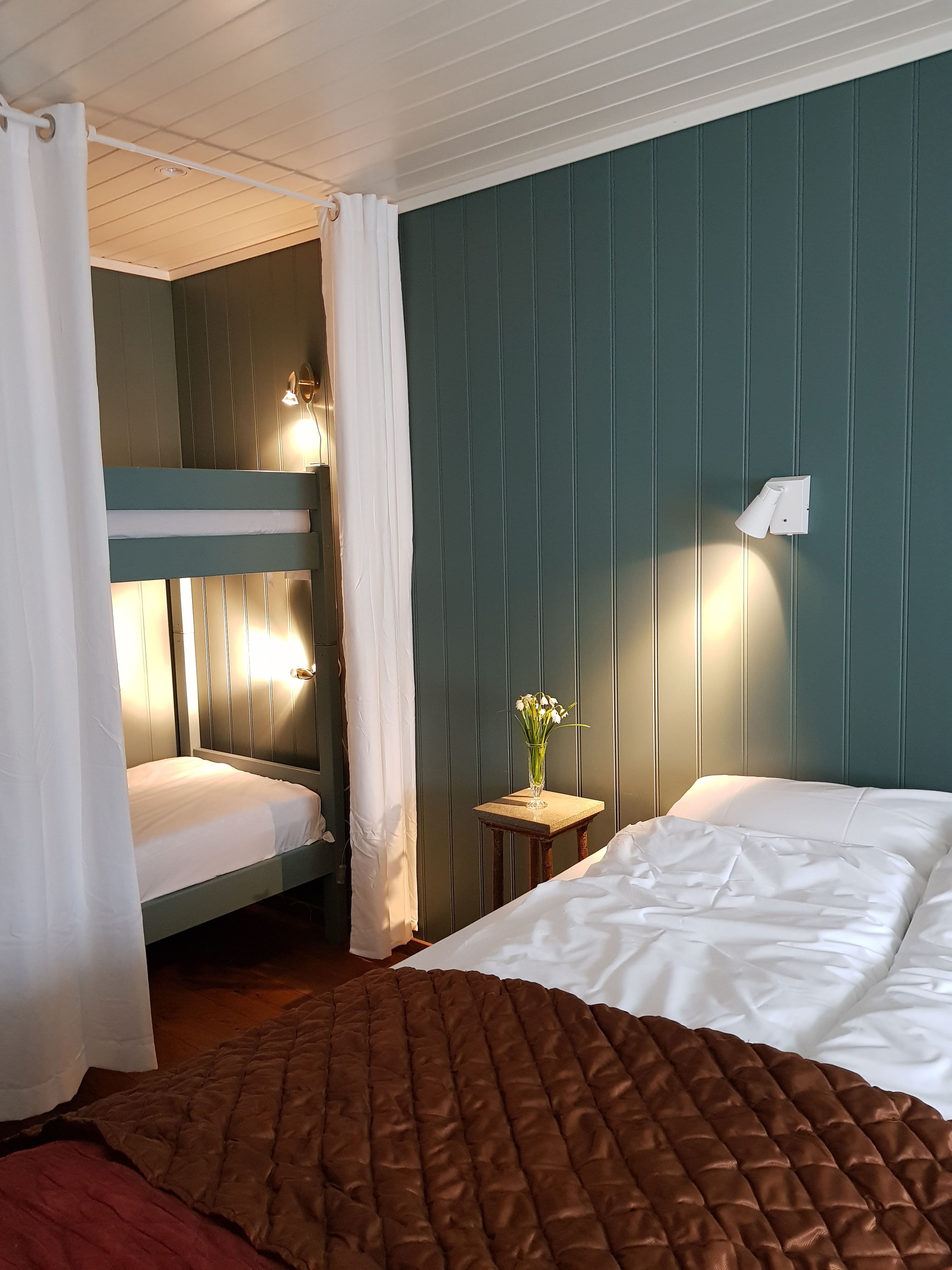 Orheimstunet in Nordfjord - Cozy bedroom with a bunk bed behind white curtains, a double bed, small nightstand with flowers, and wall lights..