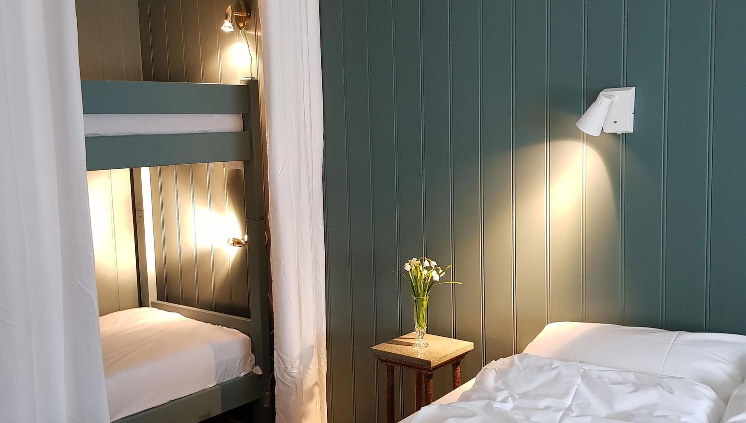 Orheimstunet in Nordfjord - Cozy bedroom with a bunk bed behind white curtains, a double bed, small nightstand with flowers, and wall lights..