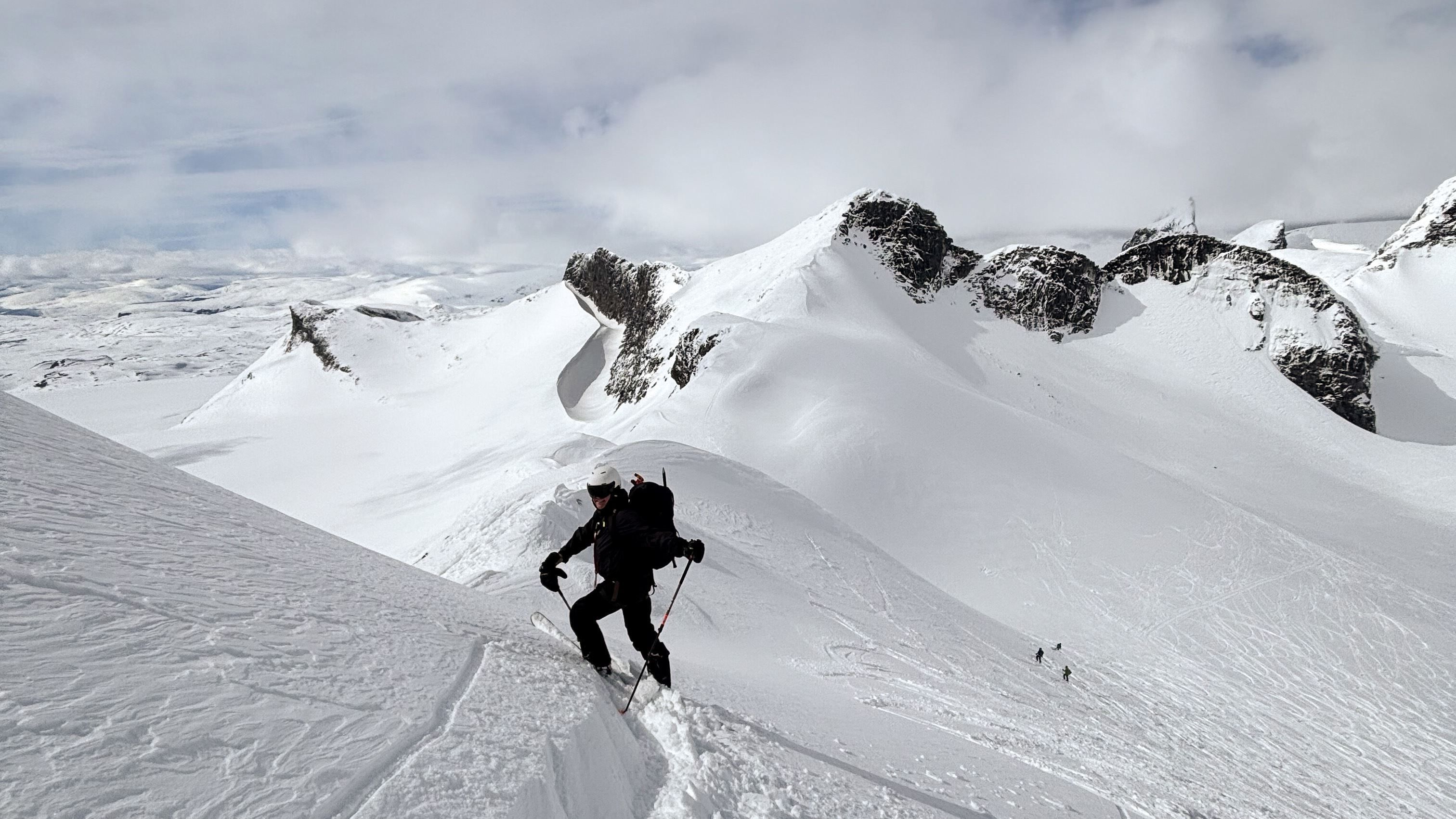 A skier with poles and a backpack climbs a narrow snowy ridge, with rugged snow-covered peaks and clouds behind..