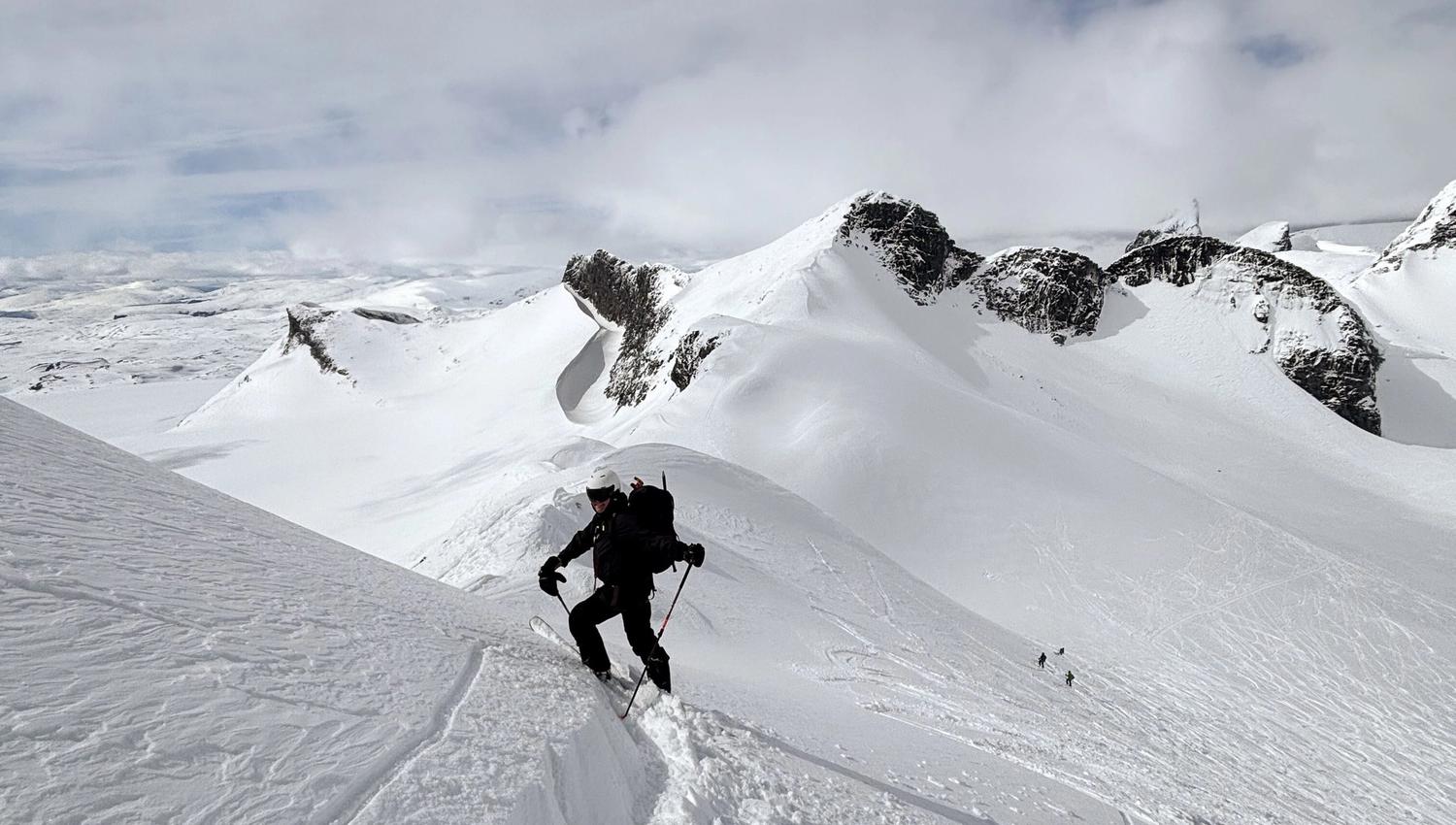 A skier with poles and a backpack climbs a narrow snowy ridge, with rugged snow-covered peaks and clouds behind..