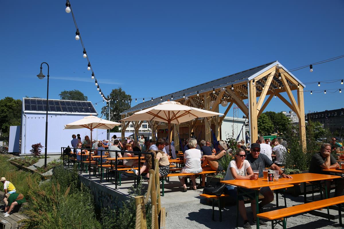 Sommerlig uteservering med mange gjester ved trebord og pergola.
