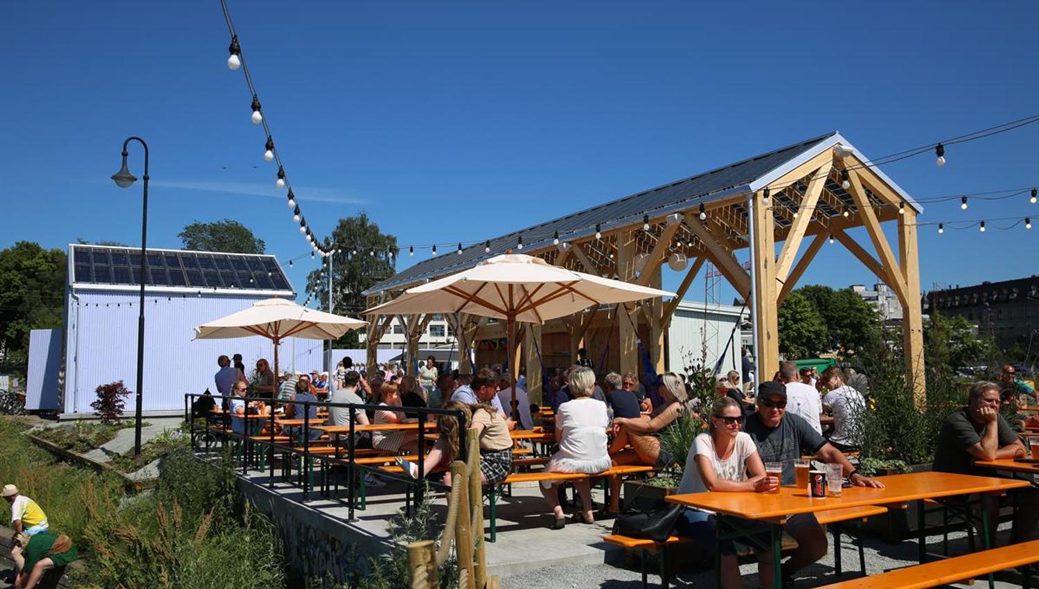 Sommerlig uteservering med mange gjester ved trebord og pergola.