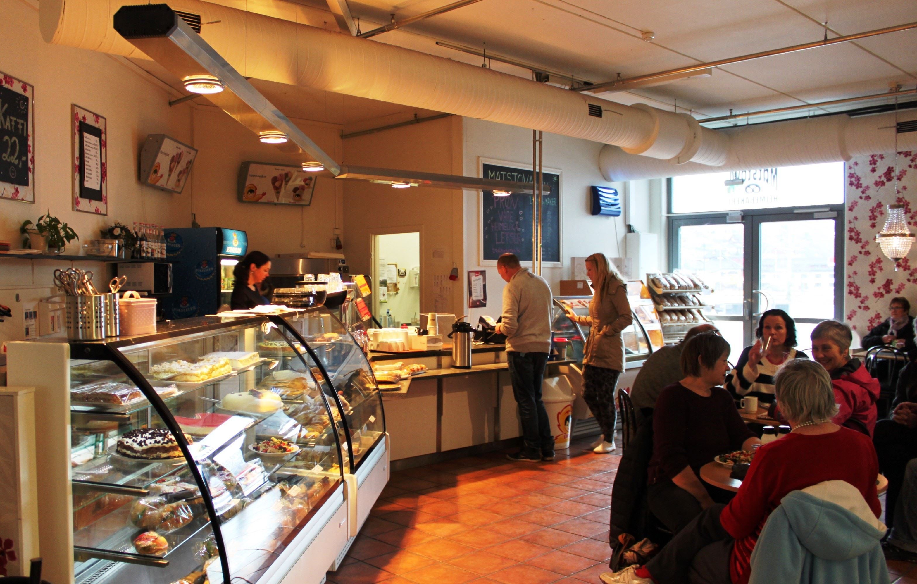 Inside a bakery cafe with glass pastry cases, customers at the counter, and people seated at tables near large windows.