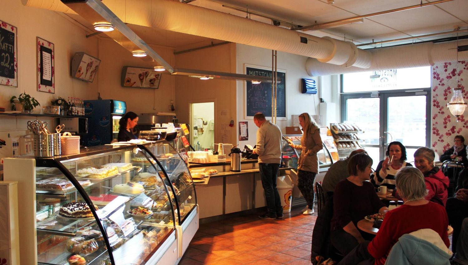 Inside a bakery cafe with glass pastry cases, customers at the counter, and people seated at tables near large windows.