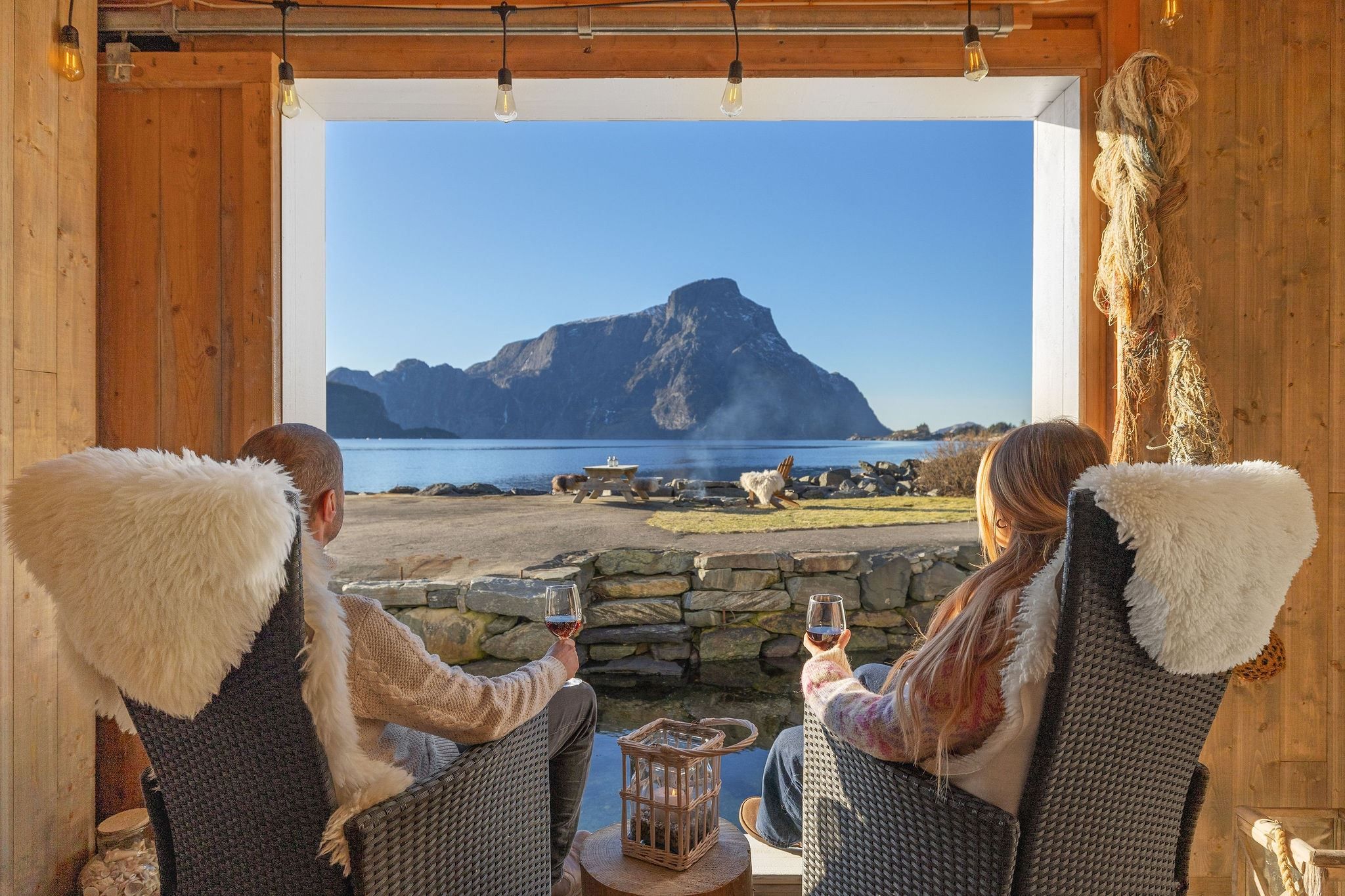 Two people sit in cozy chairs holding wine, looking out at a mountain across calm water from a wooden cabin at the Seahouse at Hornelen Panorama in Bremanger..