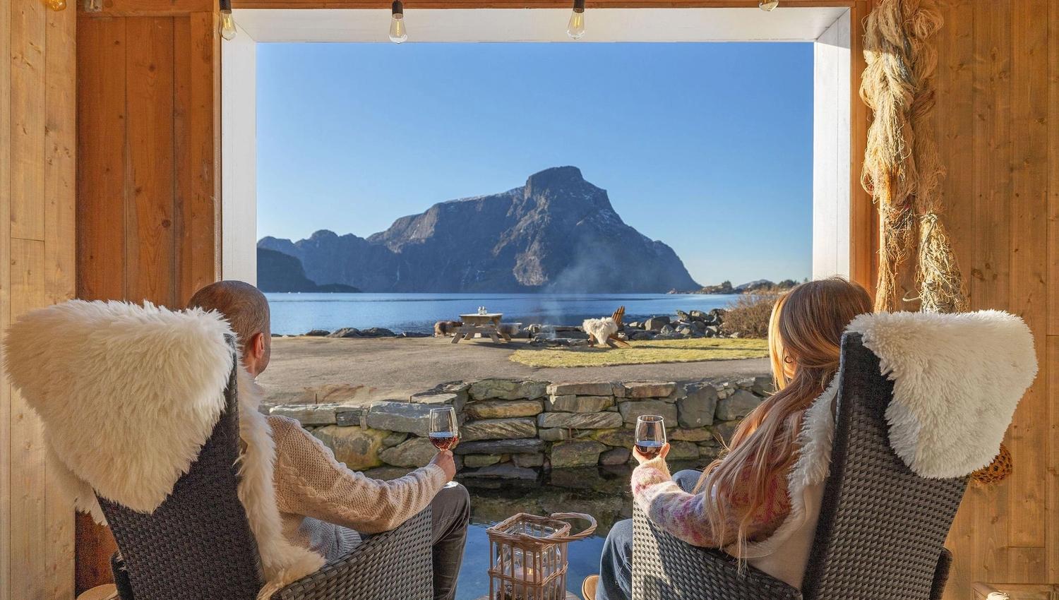 Two people sit in cozy chairs holding wine, looking out at a mountain across calm water from a wooden cabin at the Seahouse at Hornelen Panorama in Bremanger..