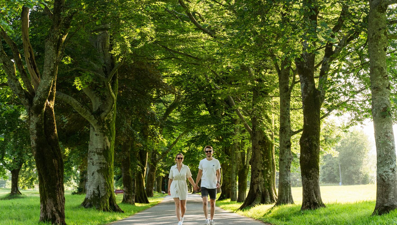 A couple walking hand in hand through an avenue of green trees at Baroniet in Rosendal on a sunny day.