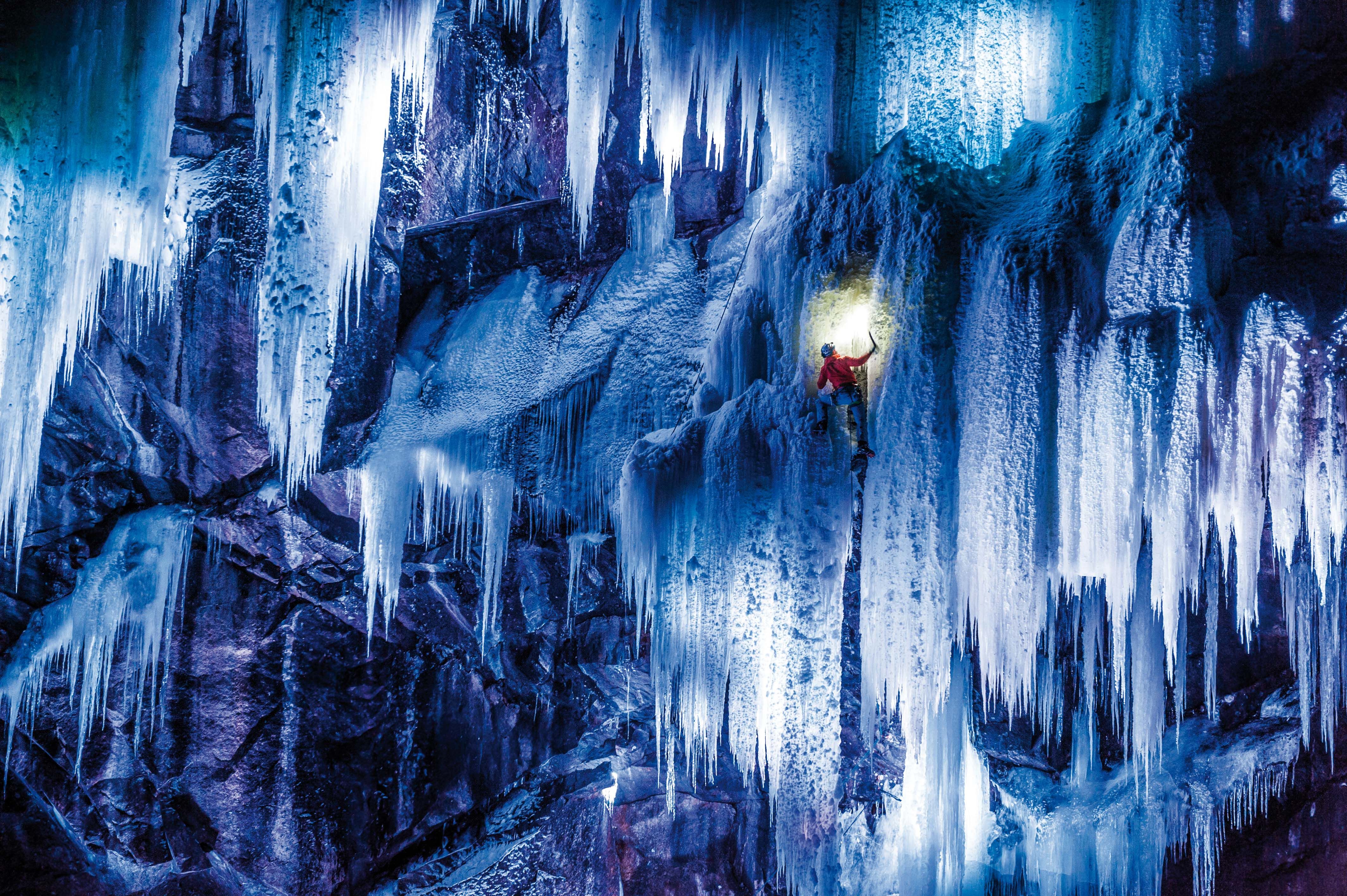 A spectacular photo of a climber climbing the ice which is lit up by blue and purple lights.