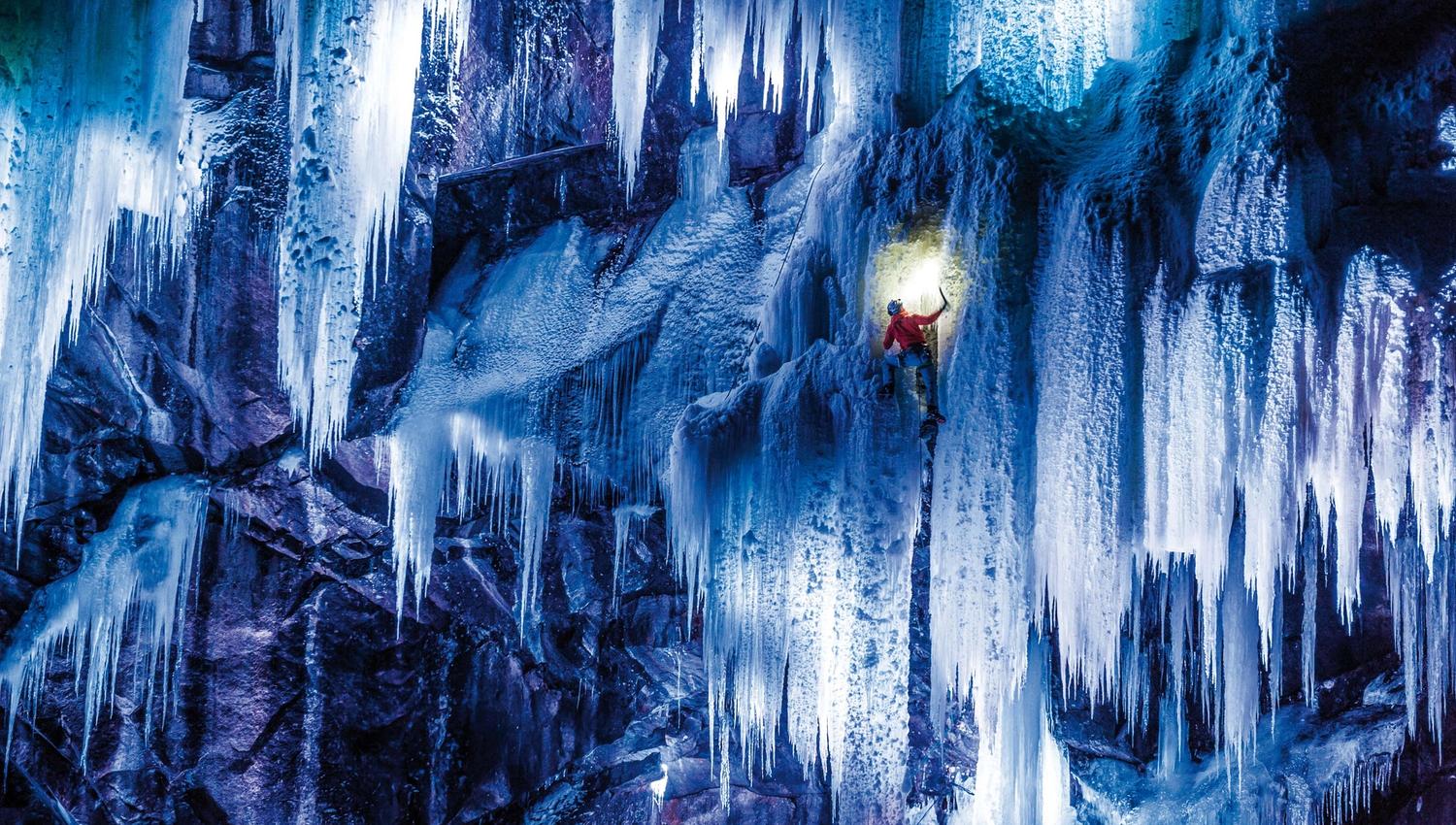 A spectacular photo of a climber climbing the ice which is lit up by blue and purple lights.