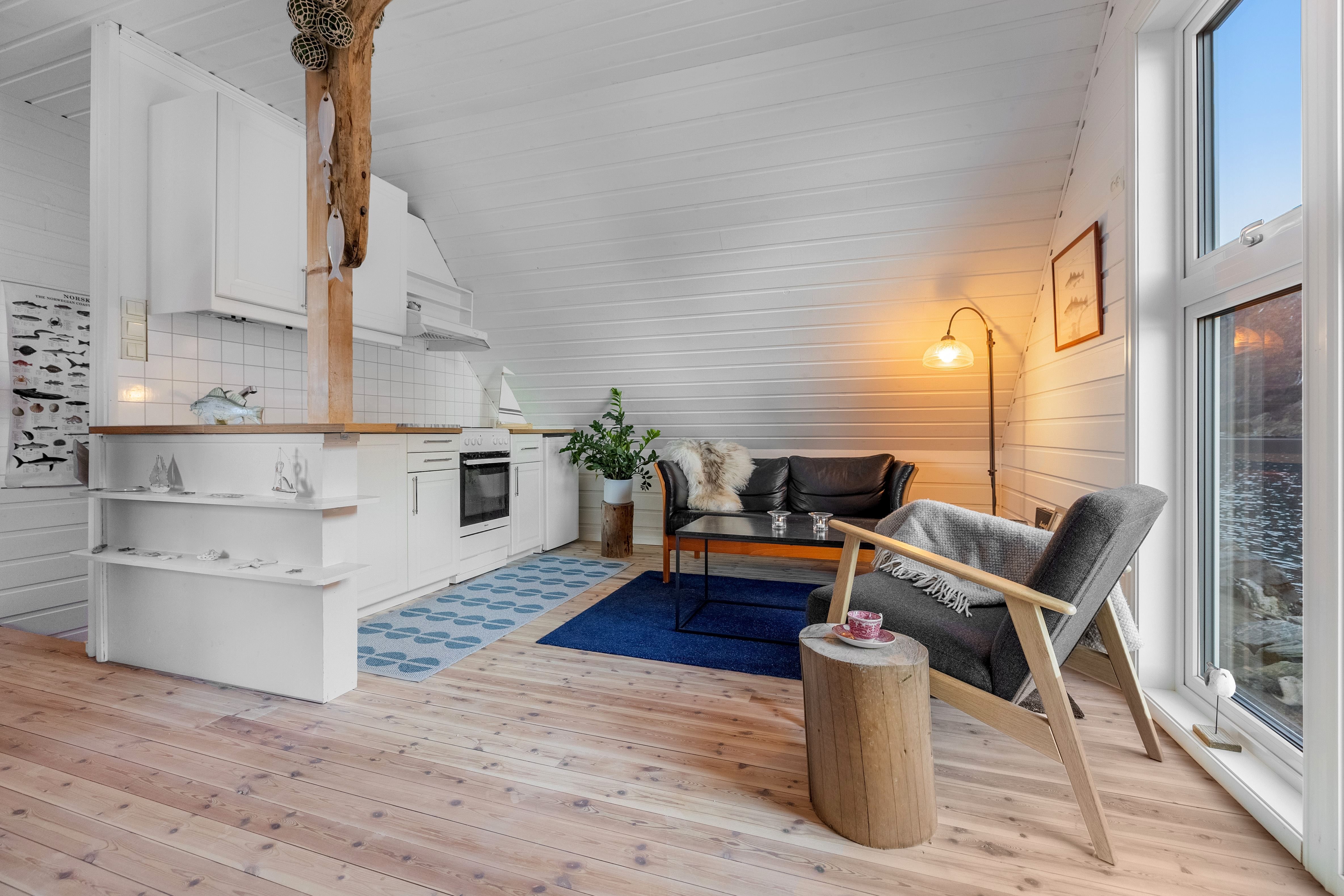 Bright wood-paneled cabin interior with white kitchen, sofa, armchair, coffee table, and large window by the sea at the Seahouse at Hornelen Panorama in Bremanger..